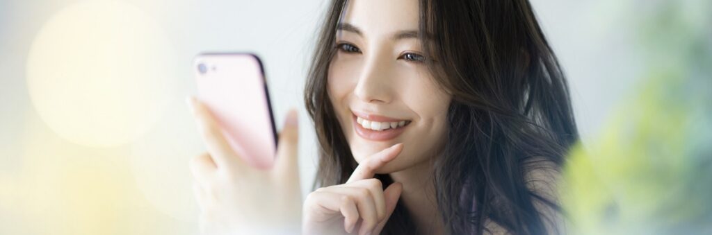 ピンクのスマートフォンを持ち、操作している人。背景は柔らかな淡い色彩でぼかされ、穏やかな雰囲気を演出しています。