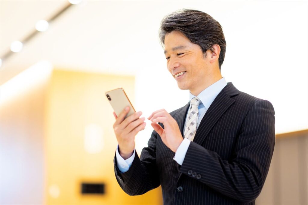 ストライプのスーツとネクタイを着用し、笑顔でスマートフォンを操作している白髪交じりのミドルエイジのビジネスマン。