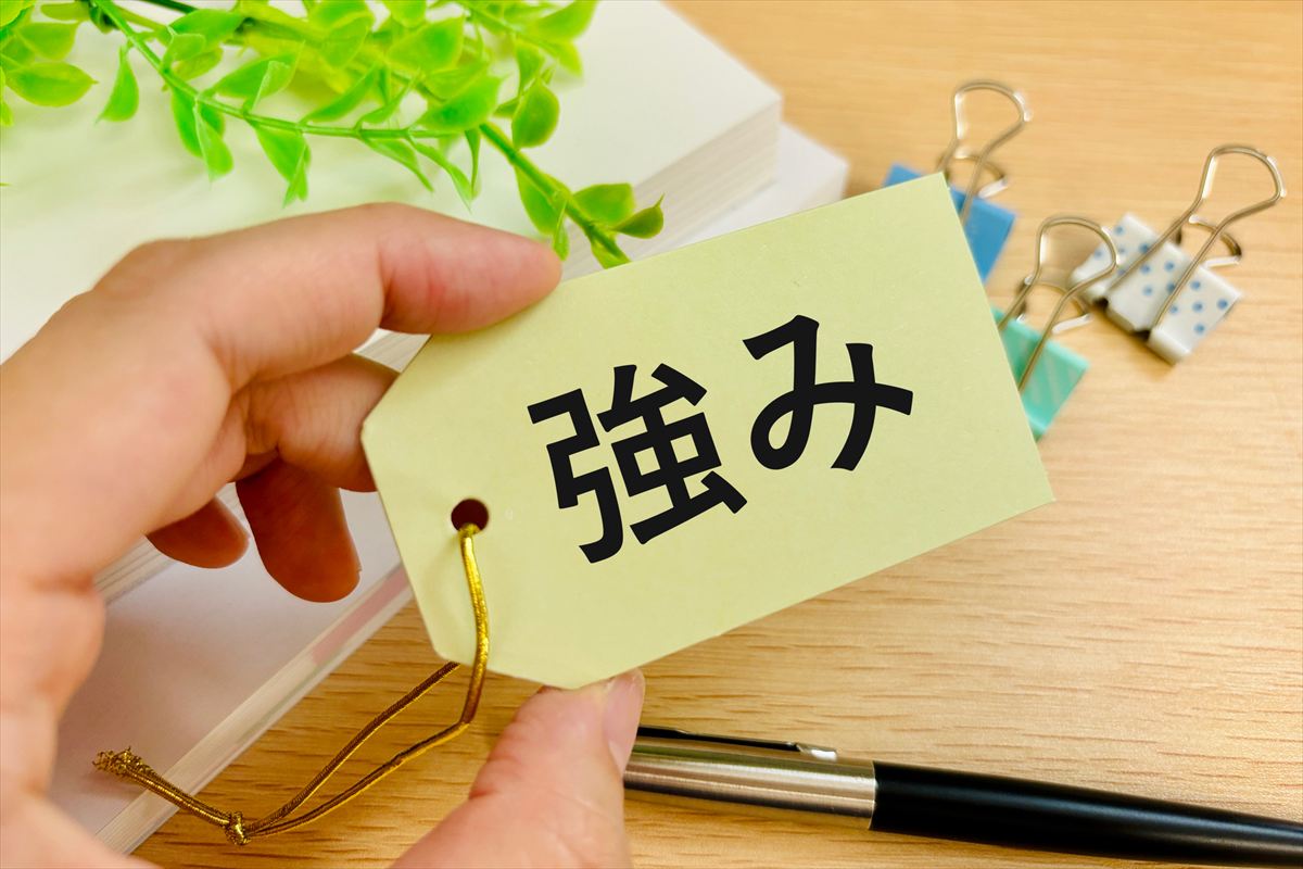 「強み」と書かれたタグを指でつまんで持っている様子。背景には書類や文具が見える。