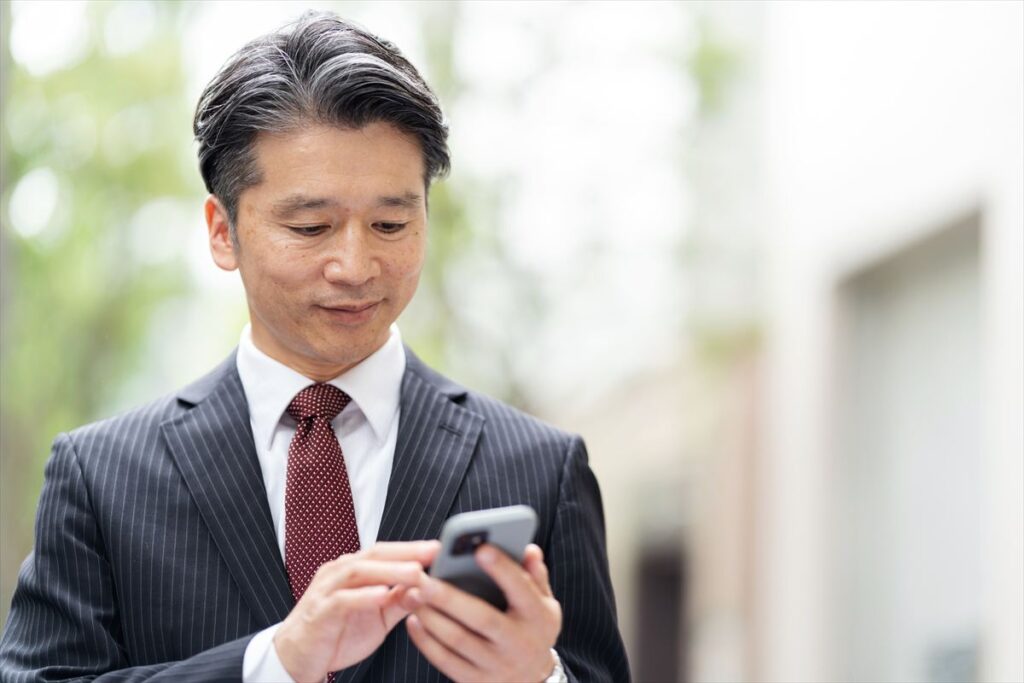 ストライプのスーツとネクタイを着用した、白髪混じりのミドルエイジの日本人ビジネスマンが、屋外でスマートフォンを操作している。