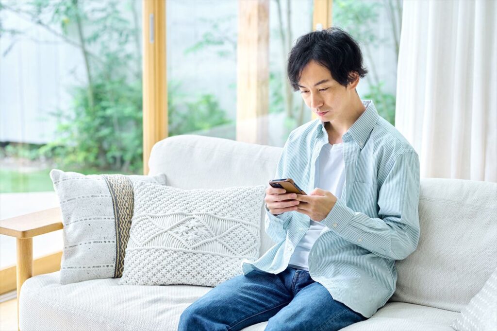 ストライプのシャツを着た若い男性が、自宅のリビングのソファに座ってスマートフォンを操作している。窓の外には緑が見える。