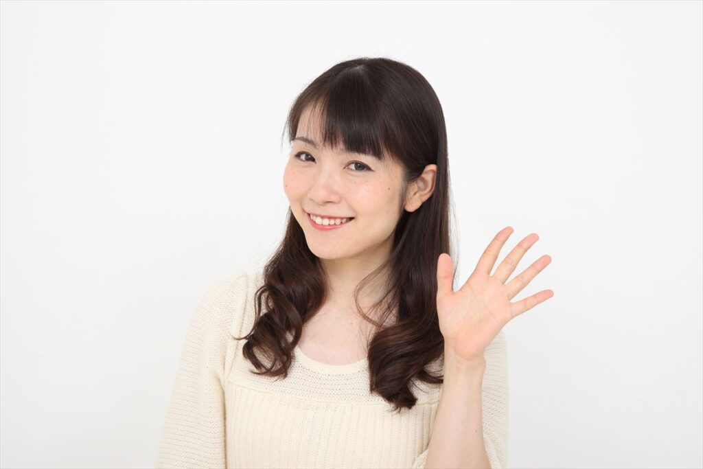 白いセーターを着た笑顔の日本人女性が、カメラに向かって手を振っている。