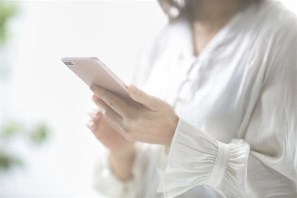 明るい日差しの中で、白いシャツを着た女性がスマートフォンを操作している様子。手元がクローズアップされ、顔は写っていない。