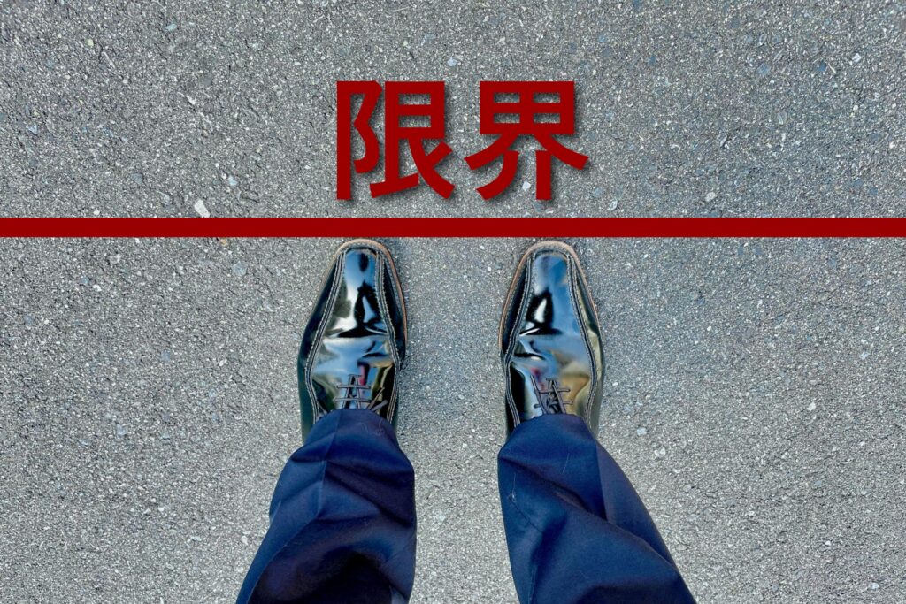 黒の革靴と紺色のスラックスを履いた人物が、アスファルトの上に立っている。足首の位置に赤い水平線が引かれ、上部には大きく「限界」の赤文字が表示されている。ビジネスや自己成長における境界線を象徴する構図。