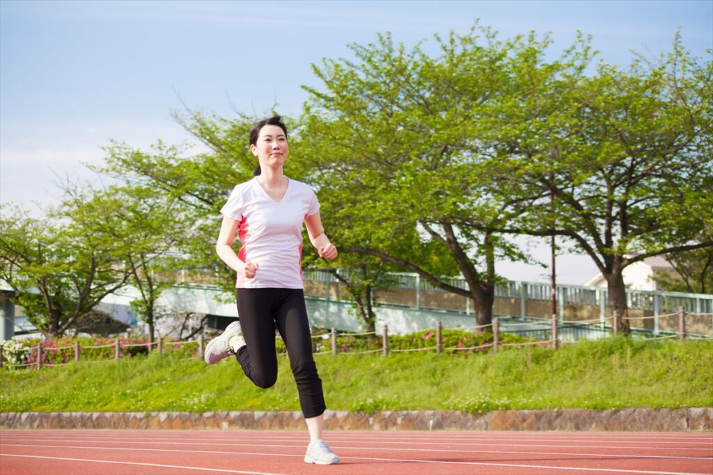 晴れた公園の赤いランニングトラックをジョギングする人物。白いTシャツに黒のパンツ、白いランニングシューズを着用し、背景には木々や歩道橋が見える。